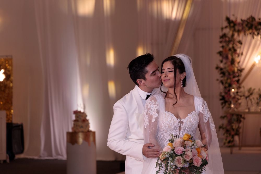 Black and white wedding photo of groom kissing bride on cheek, bride holding bouquet, elegant draped backdrop.