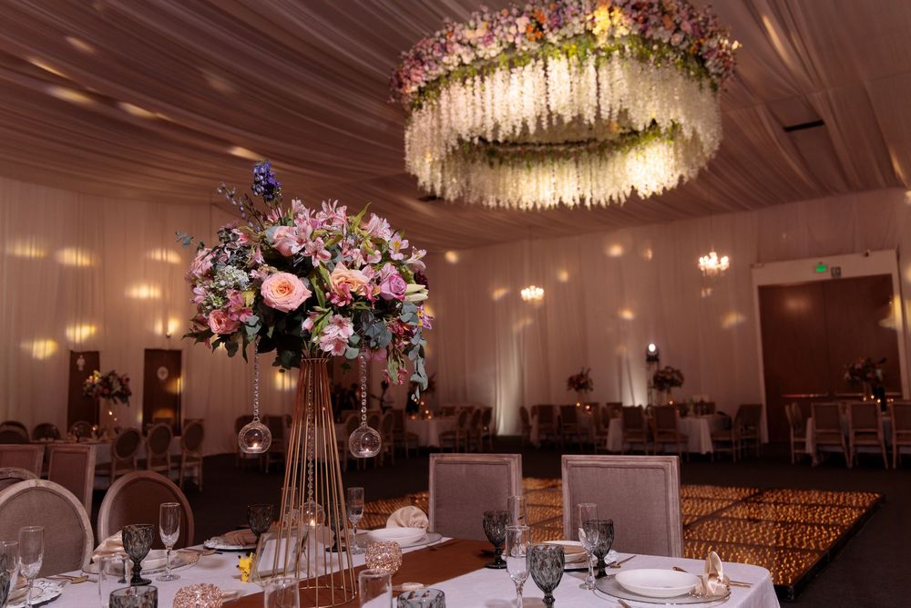 Elegant wedding reception hall with floral chandelier, tall centerpieces, and warm lighting on draped walls.