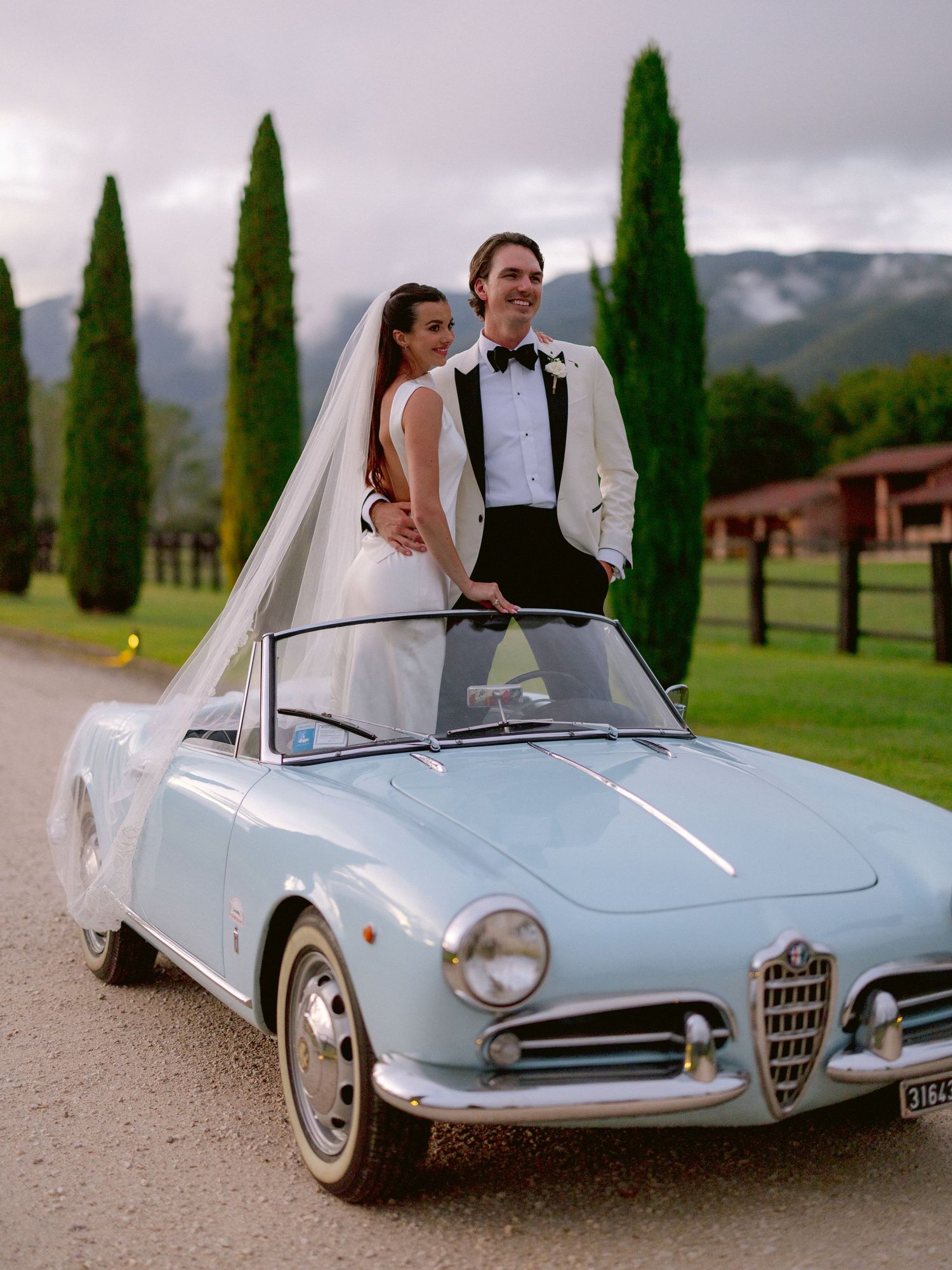 Newlyweds celebrate under a floral arch with pink and peach roses, bride in white gown, groom in white tuxedo jacket.