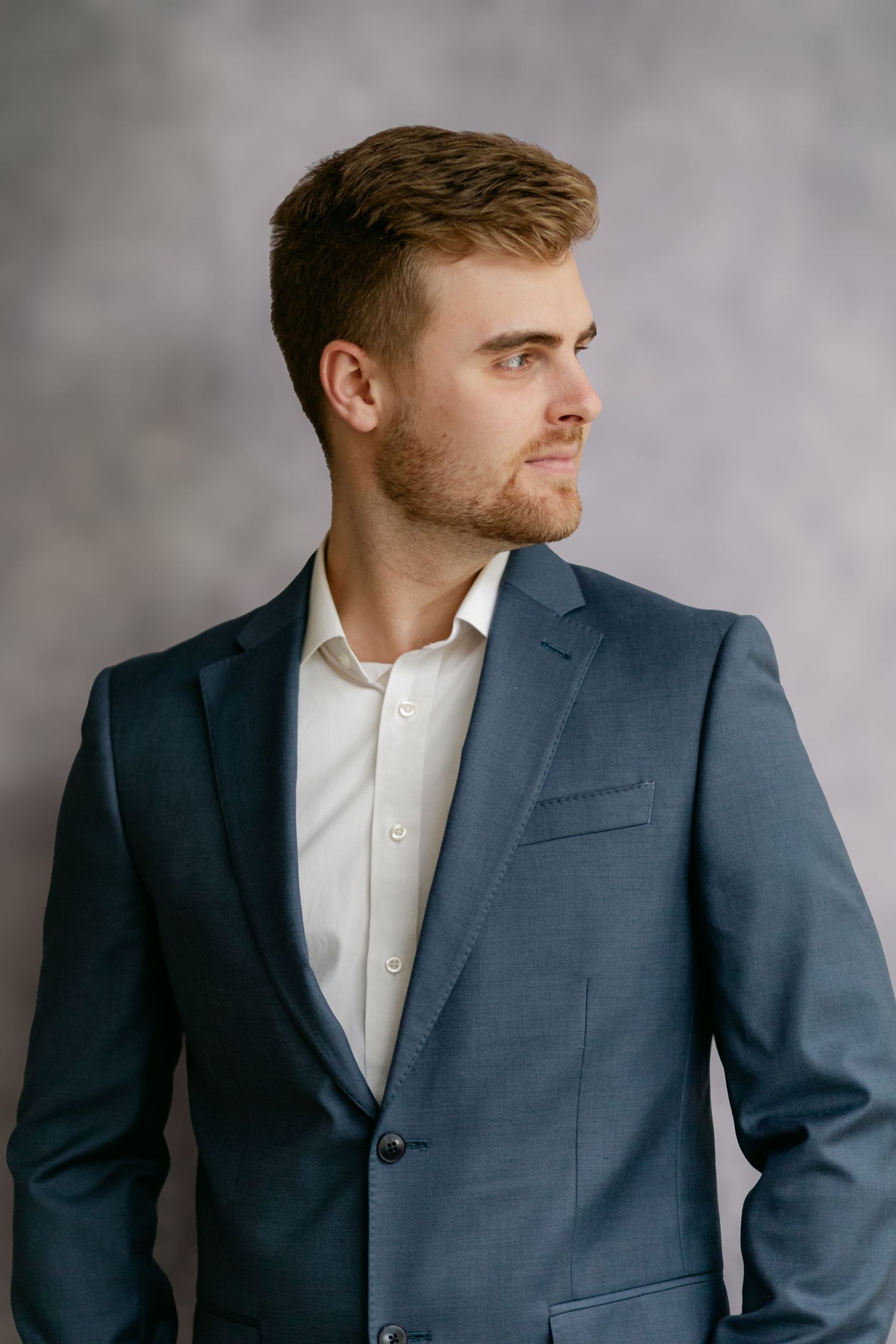 Young man with short hair in a blue suit and white shirt looking sideways in a professional studio portrait.