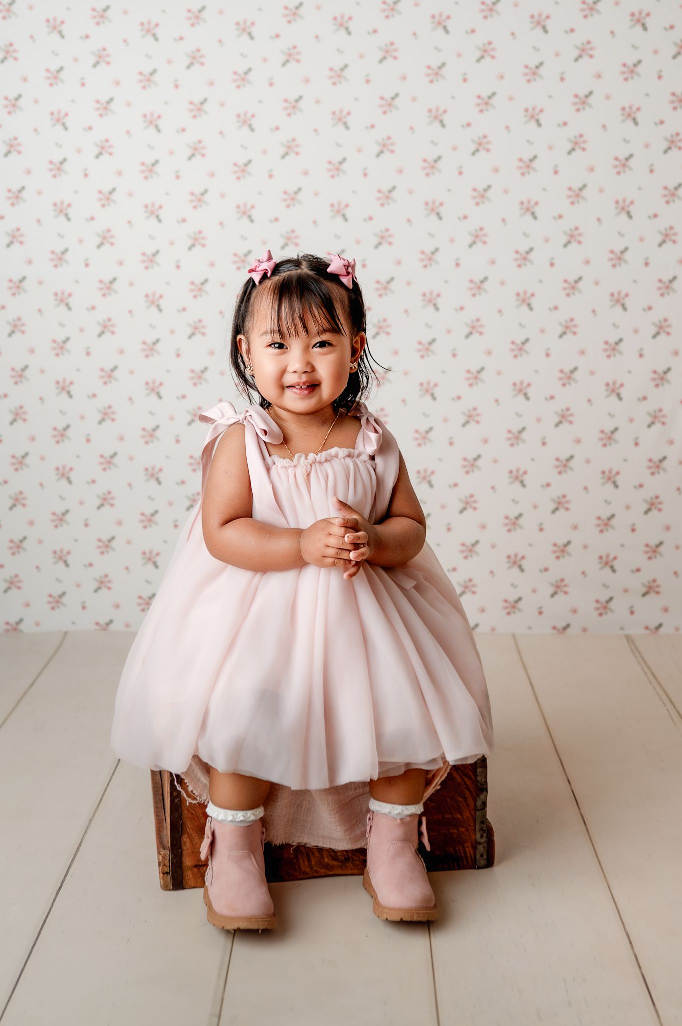 Toddler girl in pink tulle dress and boots sits on a wooden box before a floral wallpaper backdrop.
