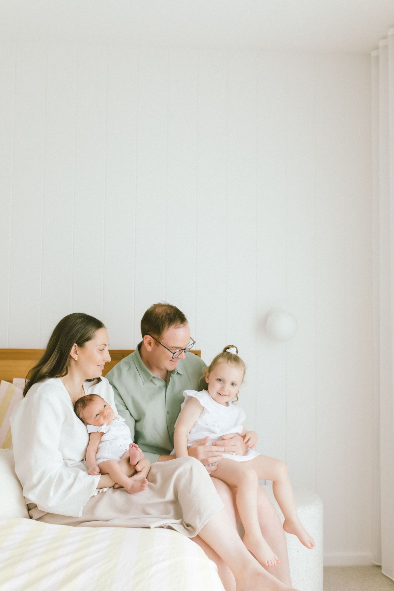 A happy family of four sits on a bed, with mother holding a newborn and father sitting beside a toddler girl.
