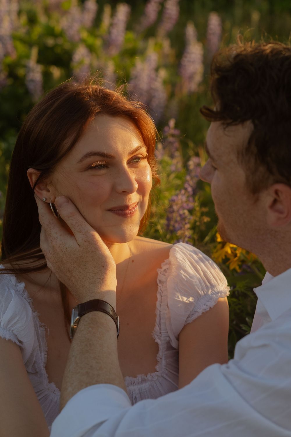 Man tenderly holding womans face while sitting among purple and yellow wildflowers at golden hour.