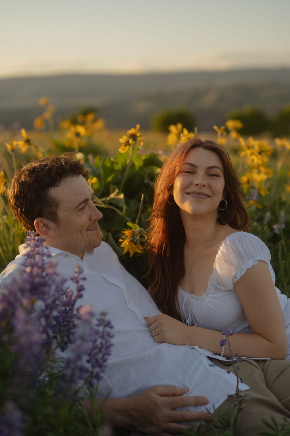 A couple sits together in a sunny wildflower field with purple lupine and yellow blooms, smiling warmly at golden hour.