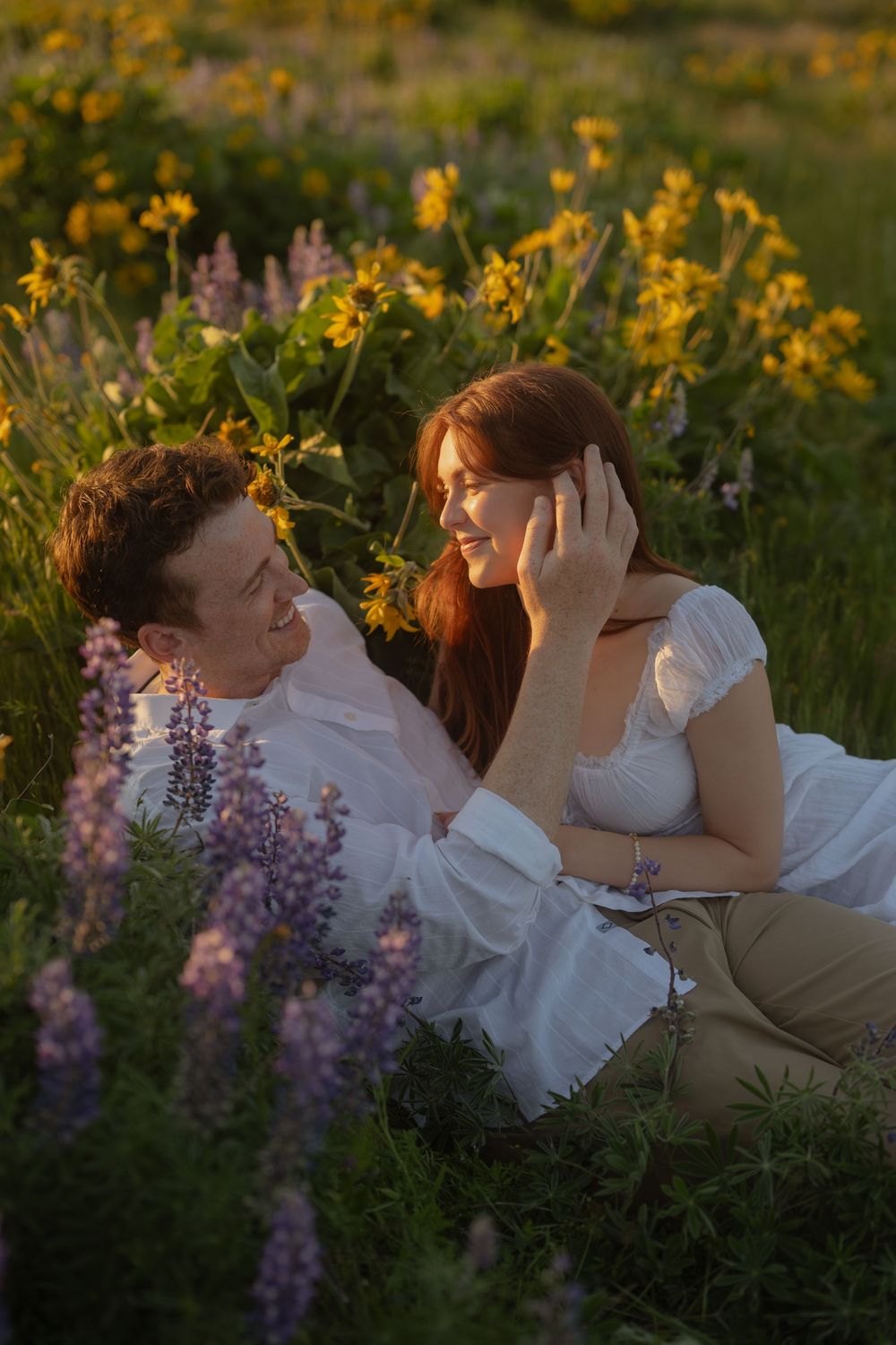 Loving couple lying together in a field of purple lupine and yellow wildflowers at sunset.