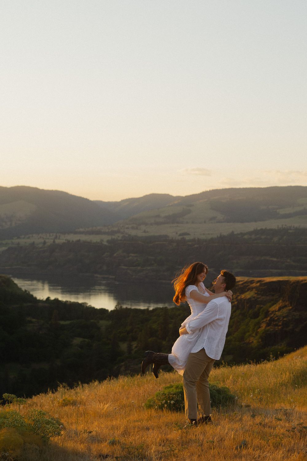 Couple embracing at golden hour on a hillside with scenic river valley and mountains in the background.