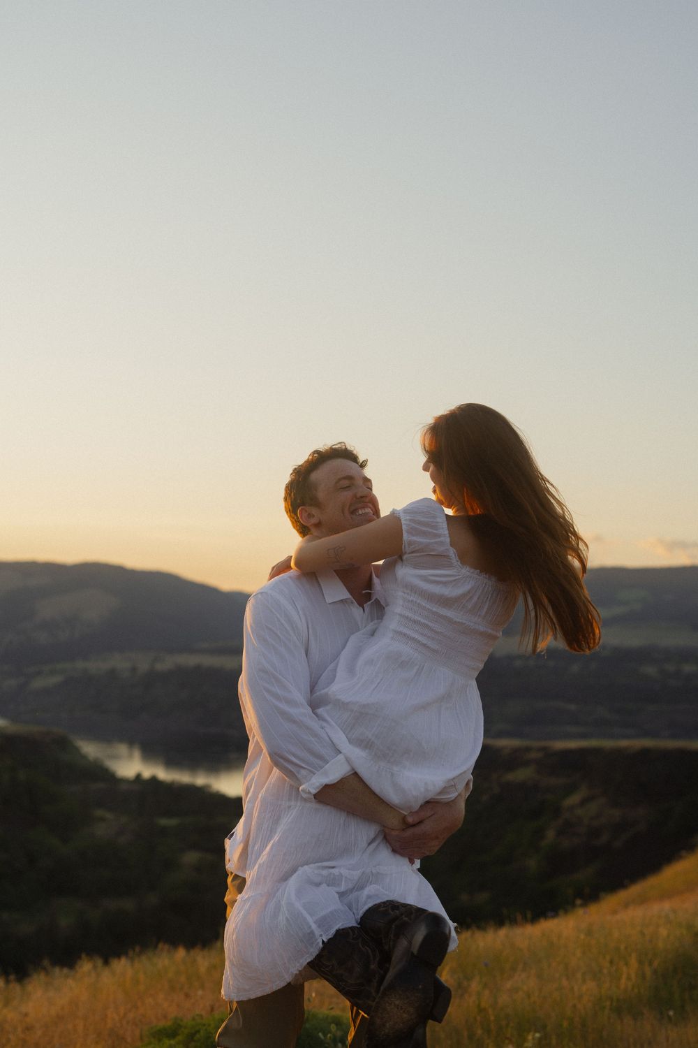 Man lifting woman in white dress at sunset with scenic river valley and mountains in background.