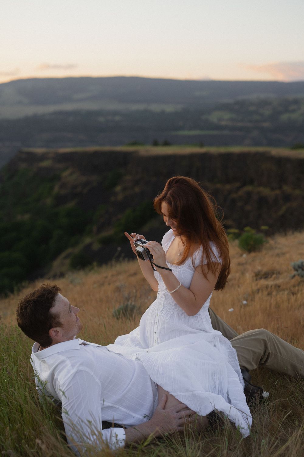 Couple in white attire relaxing on a hillside at sunset, woman holding a camera with scenic mountains in background.