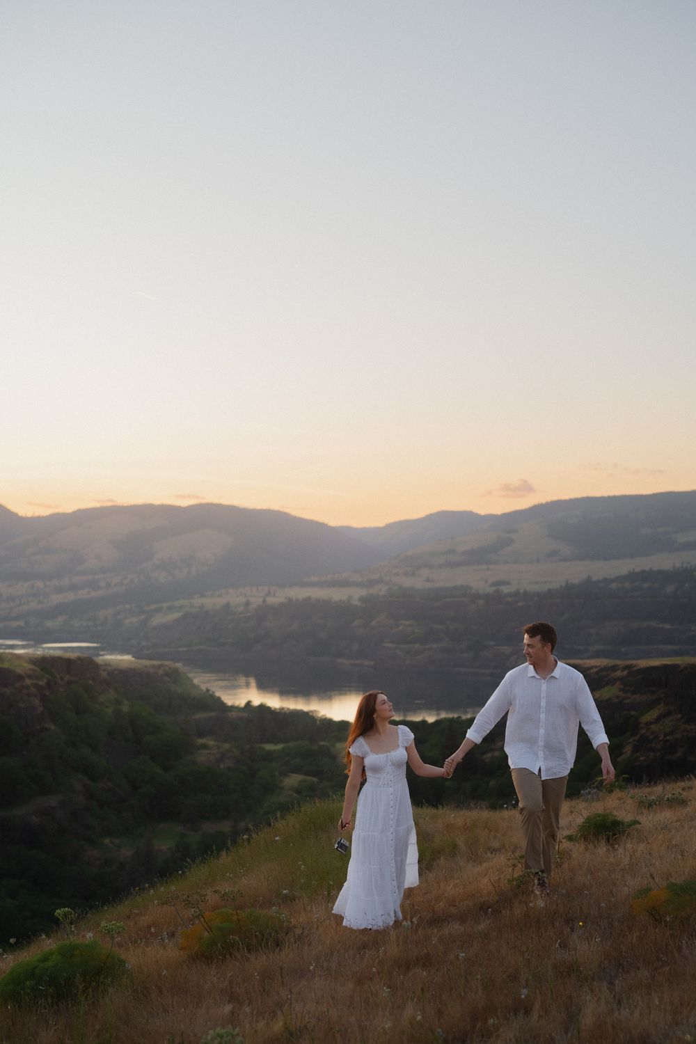 Couple holding hands at sunset on a hillside with a scenic mountain lake valley backdrop.
