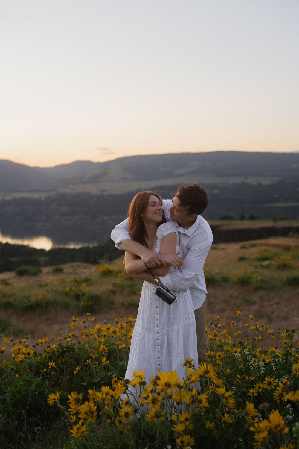 Couple embracing in a field of yellow wildflowers at sunset with mountains and lake in background.
