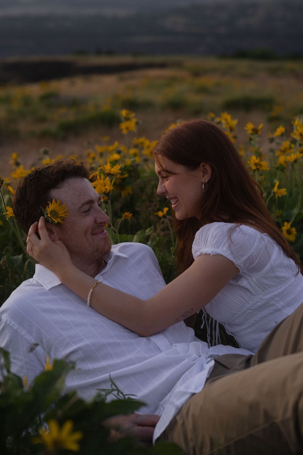 Couple sitting in yellow wildflower field at sunset, woman placing flower behind mans ear while smiling at each other.