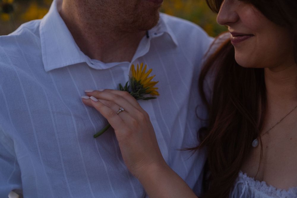 Couple posing with sunflower boutonniere, woman wearing engagement ring touching man's white striped shirt.