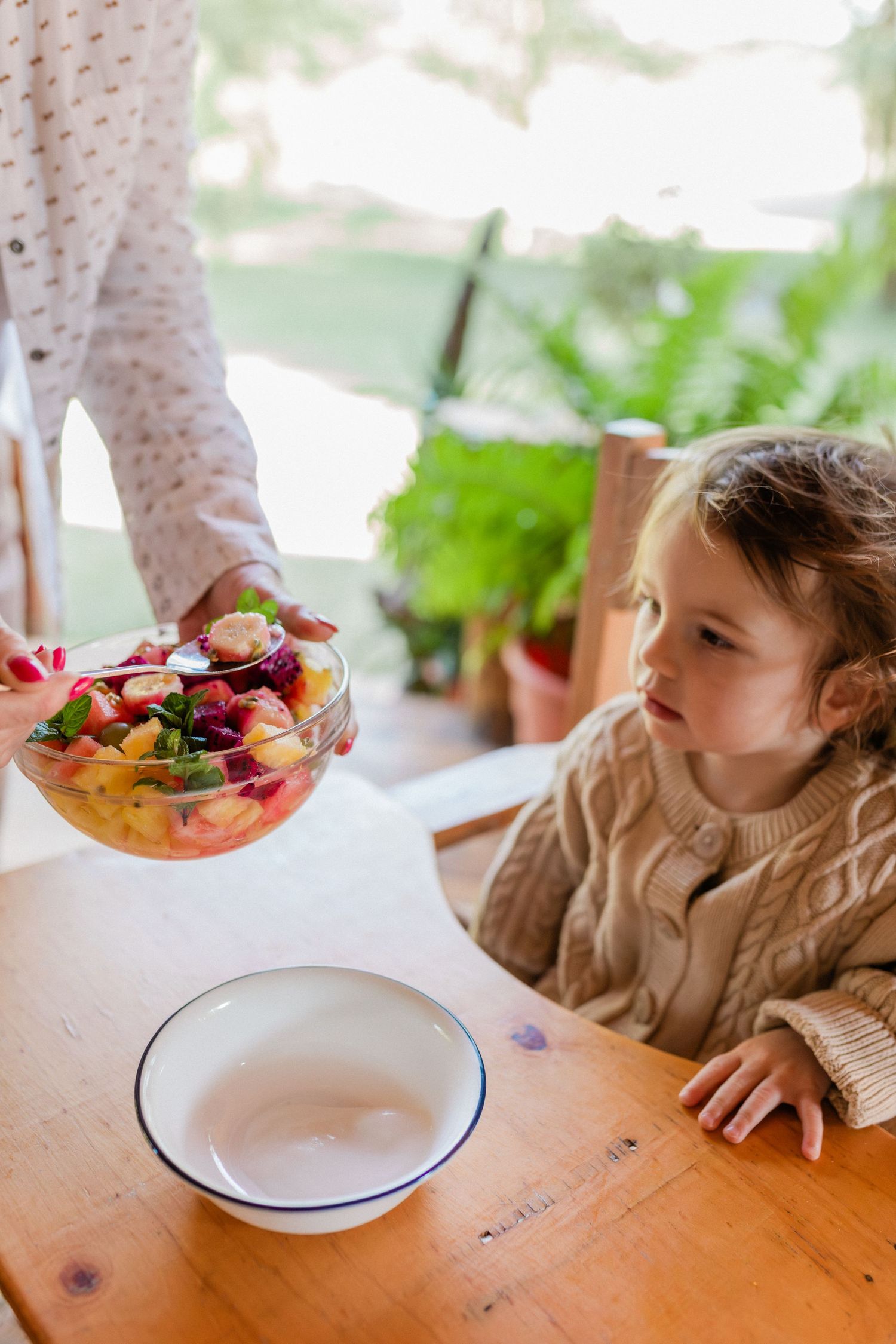 Young girl watching as an adult serves colorful fruit salad into a white bowl at a wooden dining table.