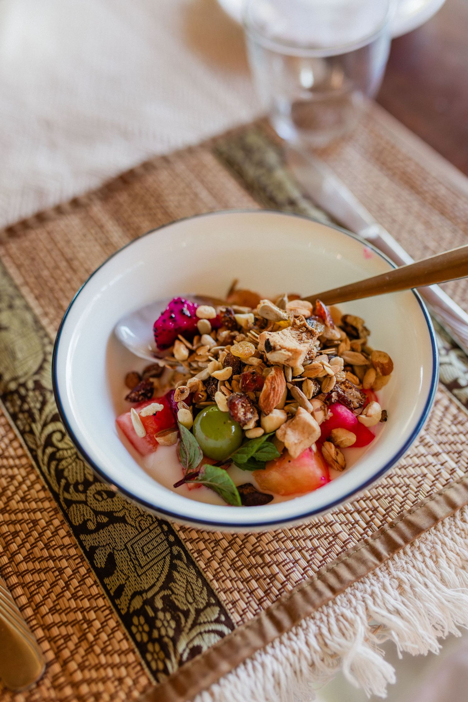 A bowl of granola with fresh fruit, nuts, and yogurt served with a golden spoon on a woven placemat.