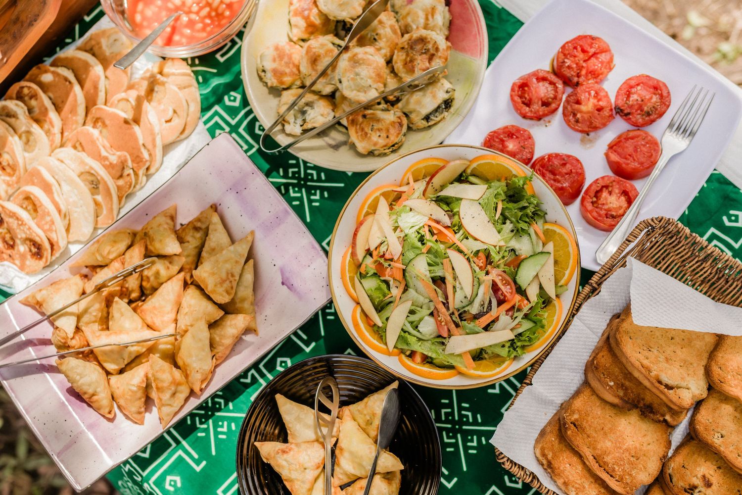 Overhead view of a colorful spread of Asian appetizers including dumplings, samosas, fresh salad, and sliced tomatoes.