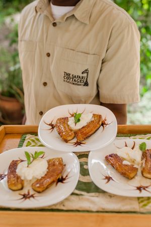 Safari Cottages server presenting three plates of caramelized bananas with cream dessert on a wooden tray.