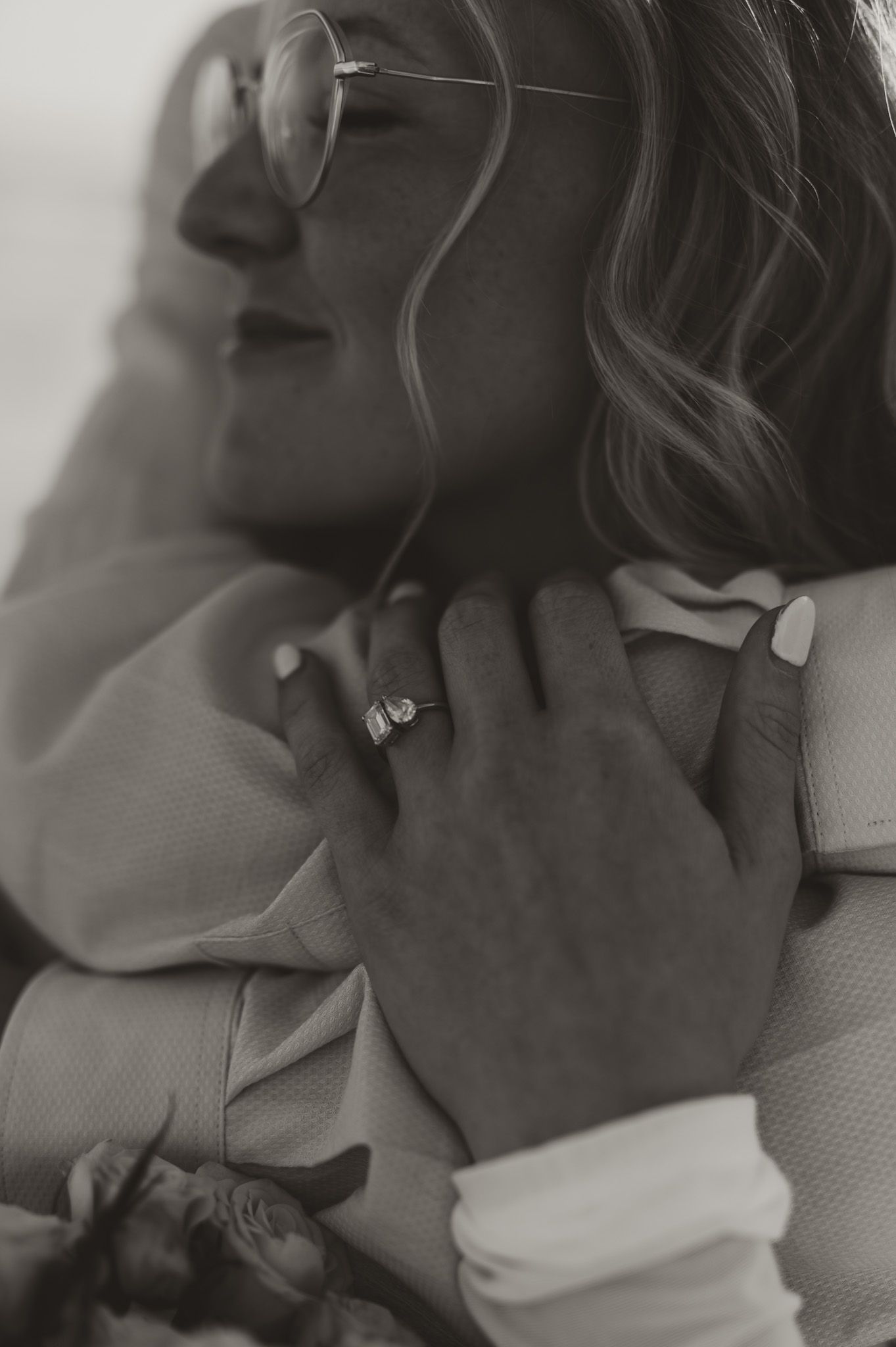 Black and white photo of a bride wearing glasses and a diamond ring embracing her groom in a bow tie.