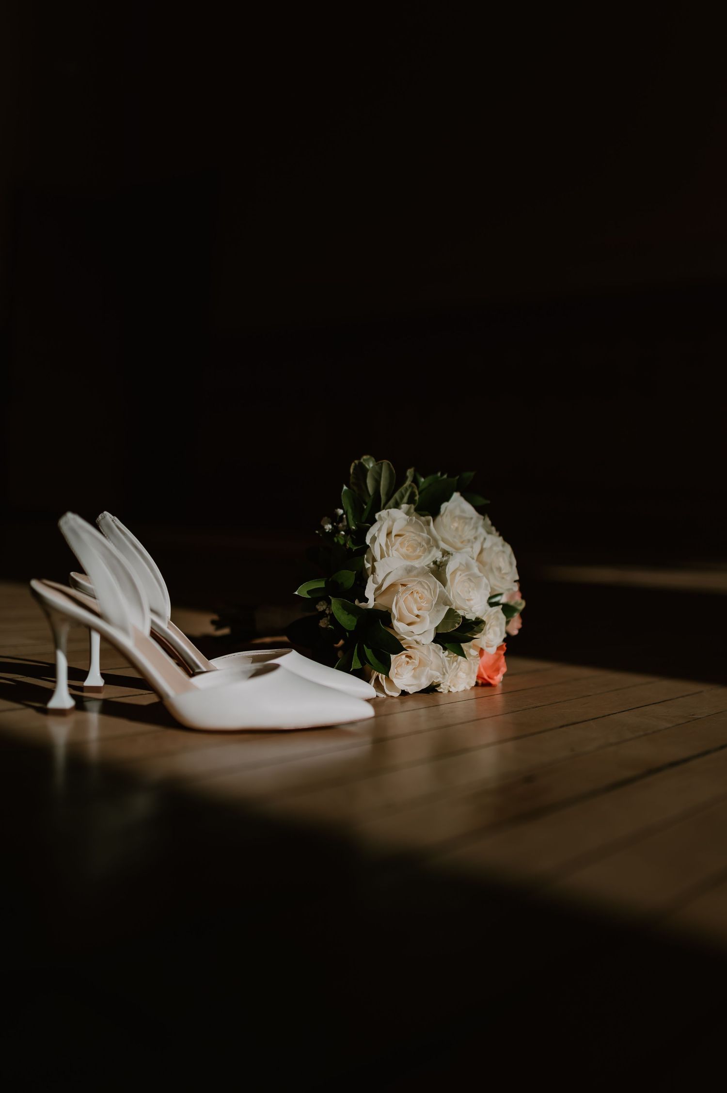 White bridal heels and a bouquet of white roses rest on a wooden floor in dramatic moody lighting during a wedding in West Bend, WI.