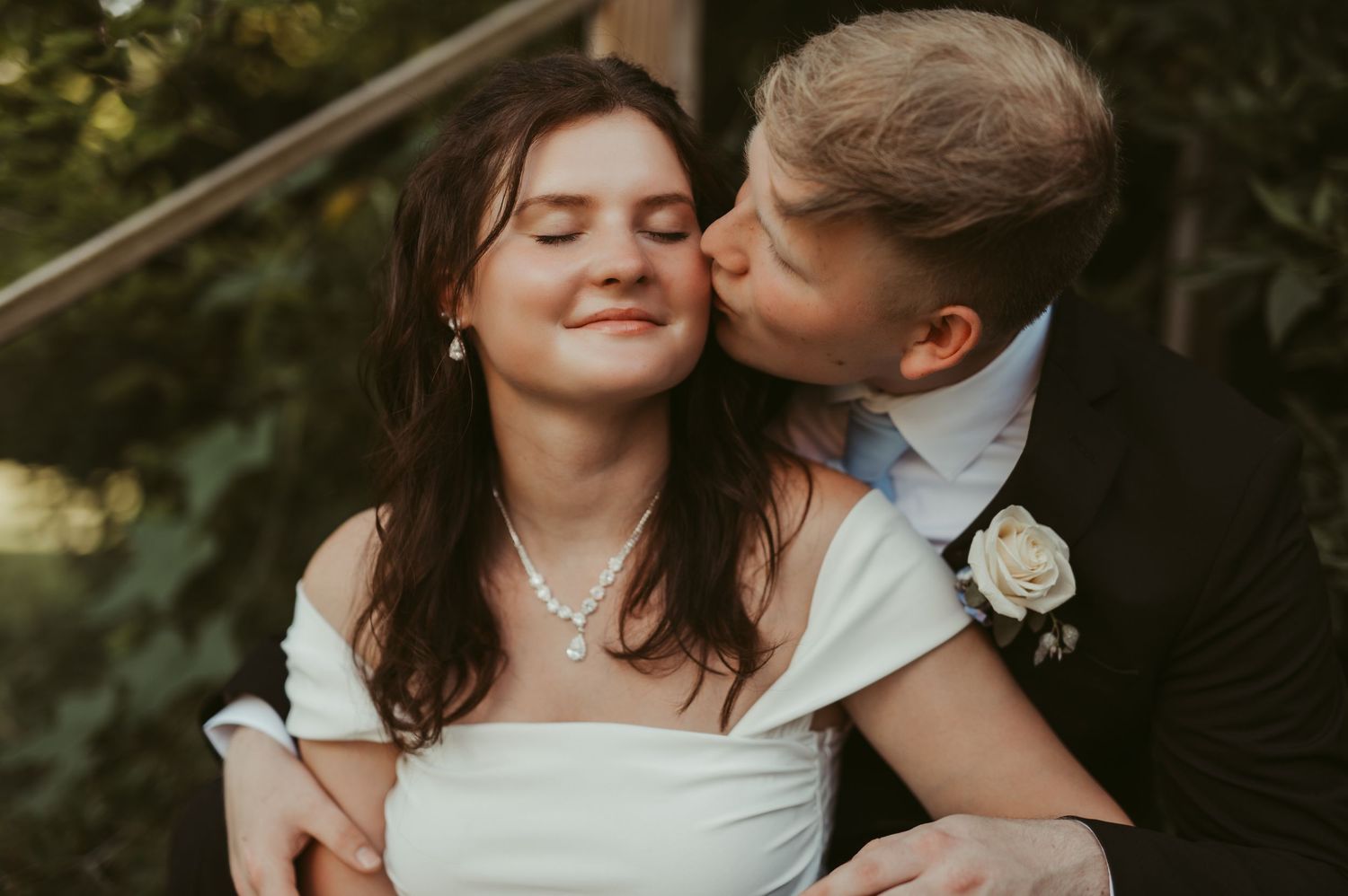 Groom kissing bride on cheek during romantic outdoor wedding portrait, bride wearing white dress and pearl necklace in Germantown, Wisconsin