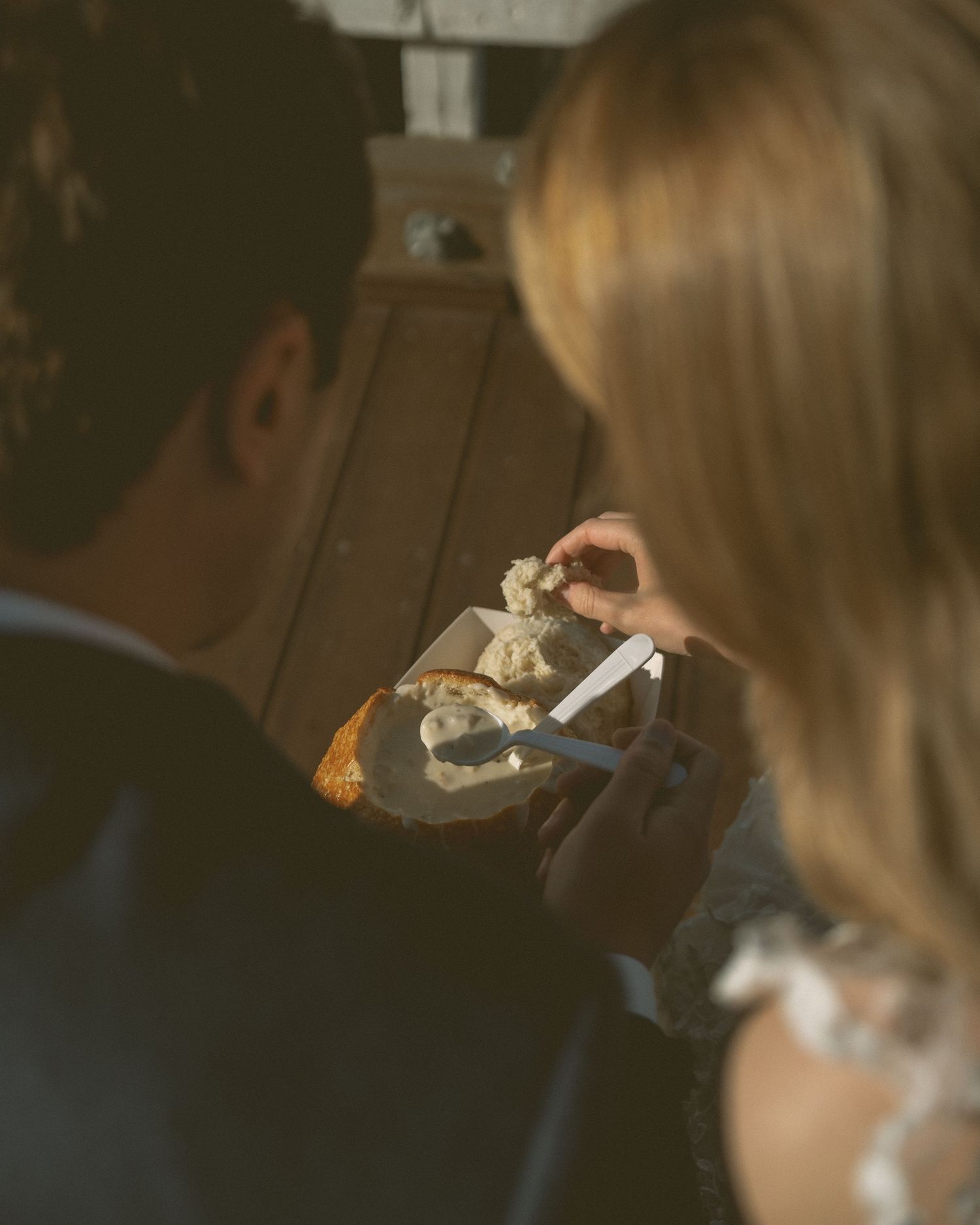 Bride and groom sharing wedding cake slice together, viewed from behind in warm sunlight.