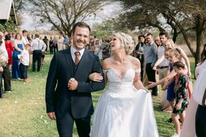 Smiling newlyweds walk through confetti shower with wedding guests celebrating in a sunny outdoor venue.