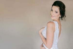 Elegant bride in a white sleeveless gown with open back, smiling over her shoulder against a neutral background.