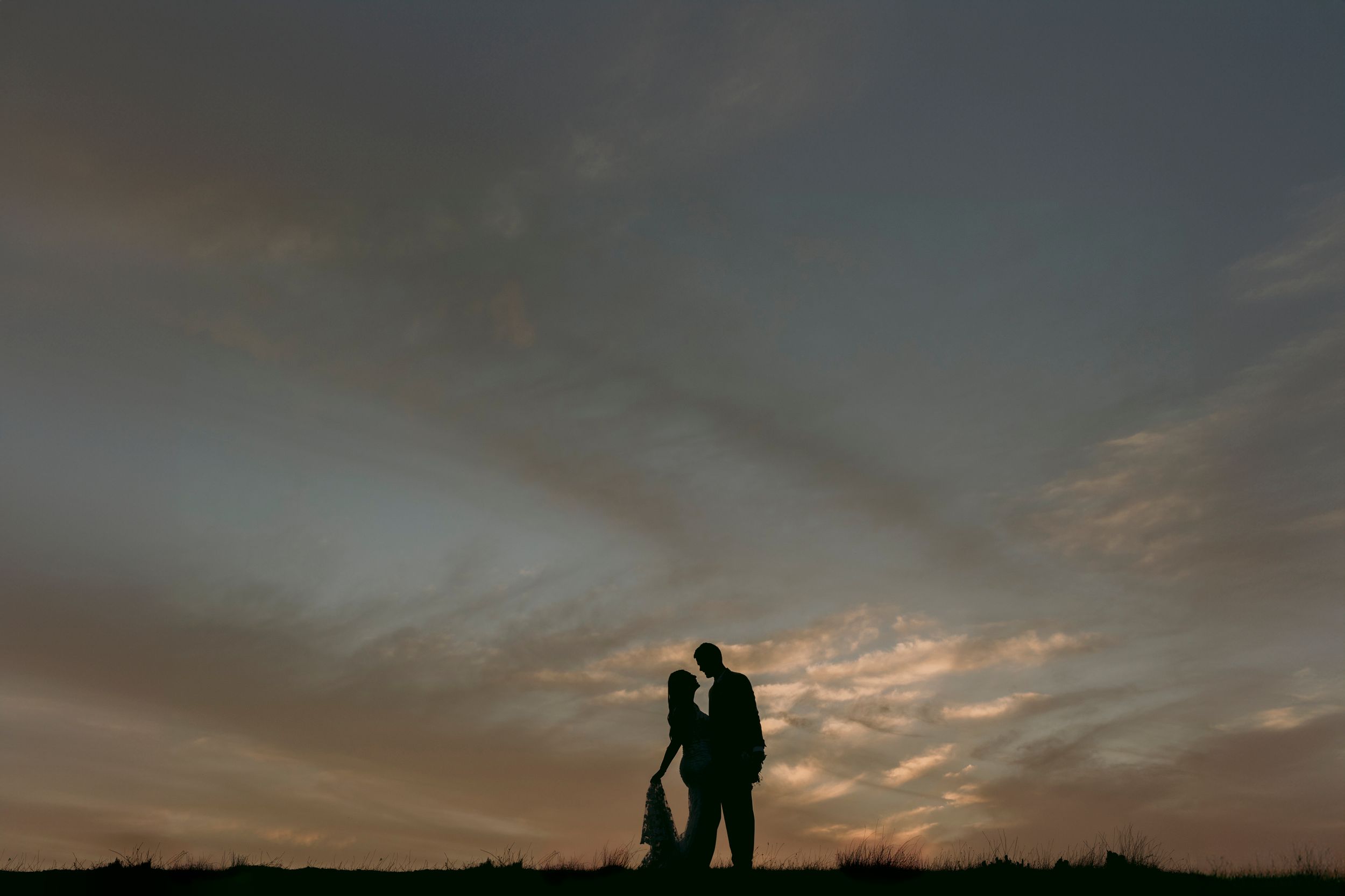 Romantic silhouette of a couple standing together against a dramatic sunset sky with clouds.