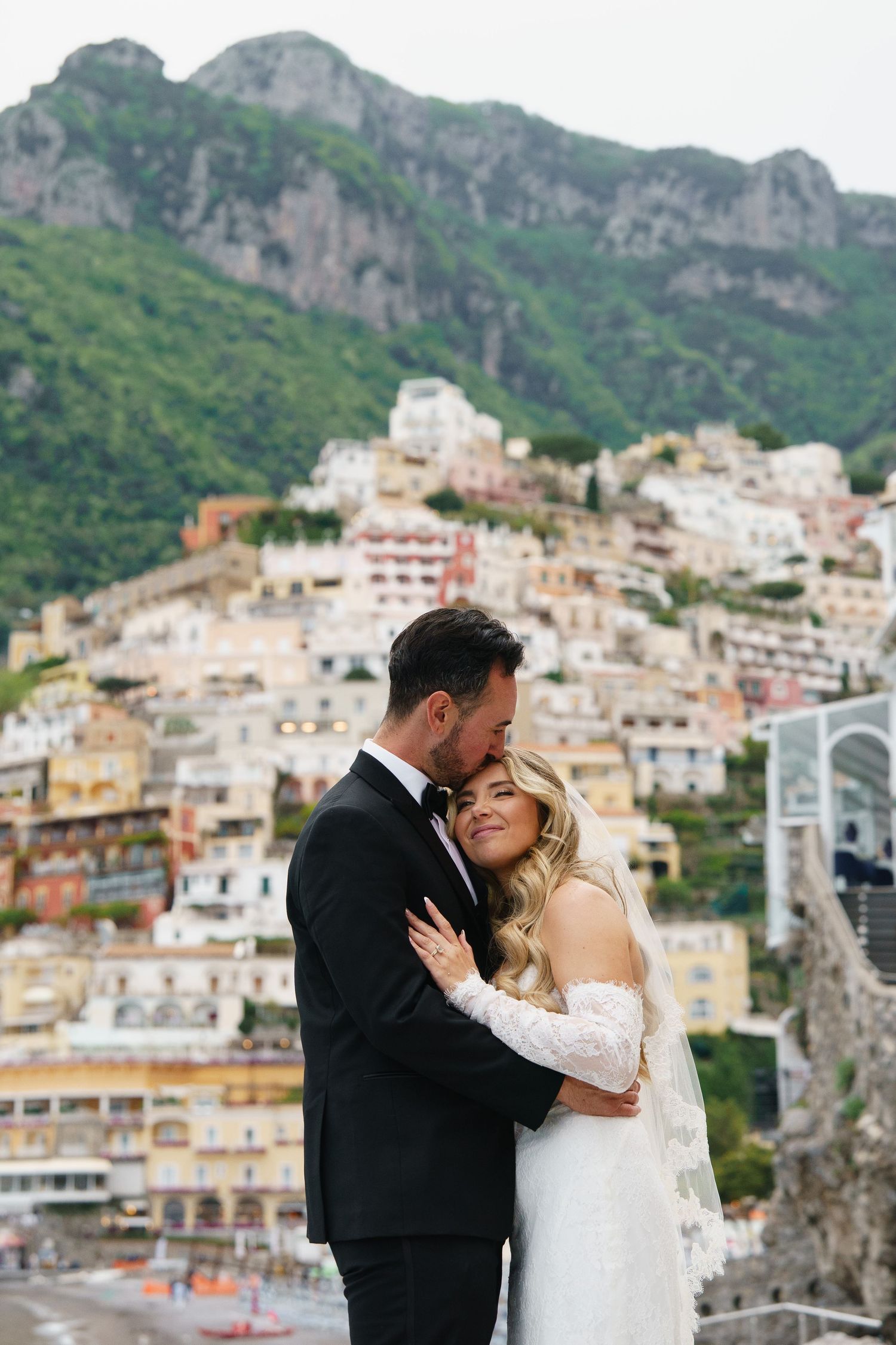 Bride and groom embrace romantically with colorful Positano hillside village backdrop on the Amalfi Coast, Italy.