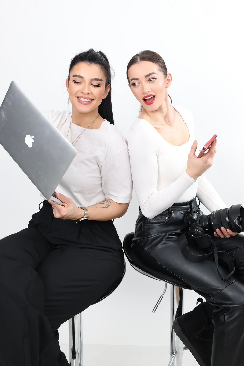 Two stylish women sitting back-to-back on chairs, one holding a MacBook laptop and the other a smartphone and camera.
