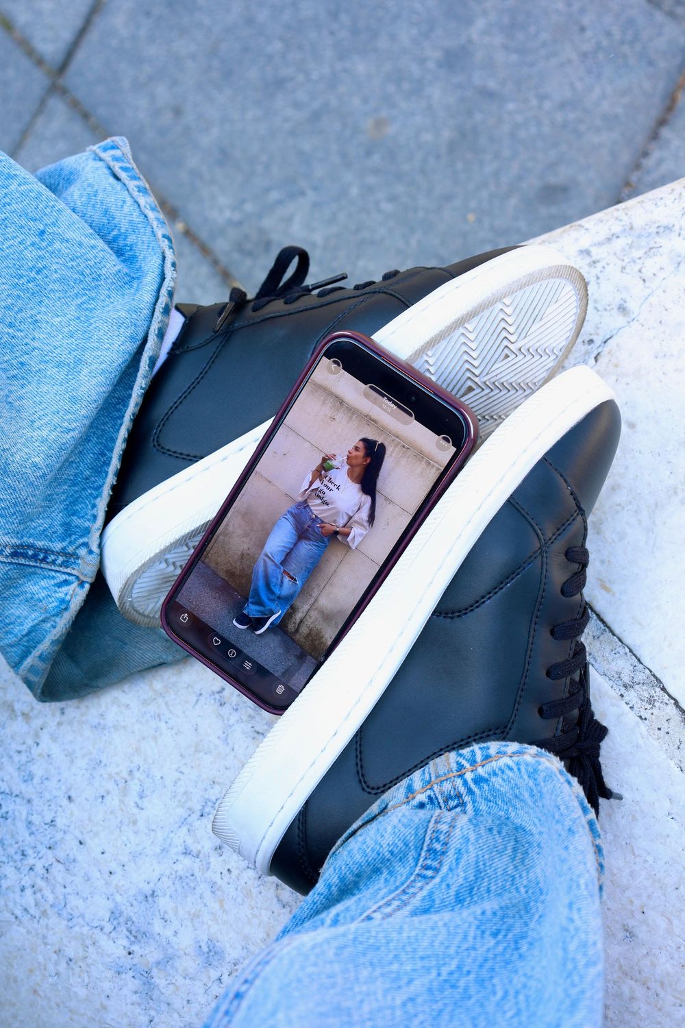 Black high-top sneakers with white soles resting on steps, with a smartphone displaying a woman holding a dog.