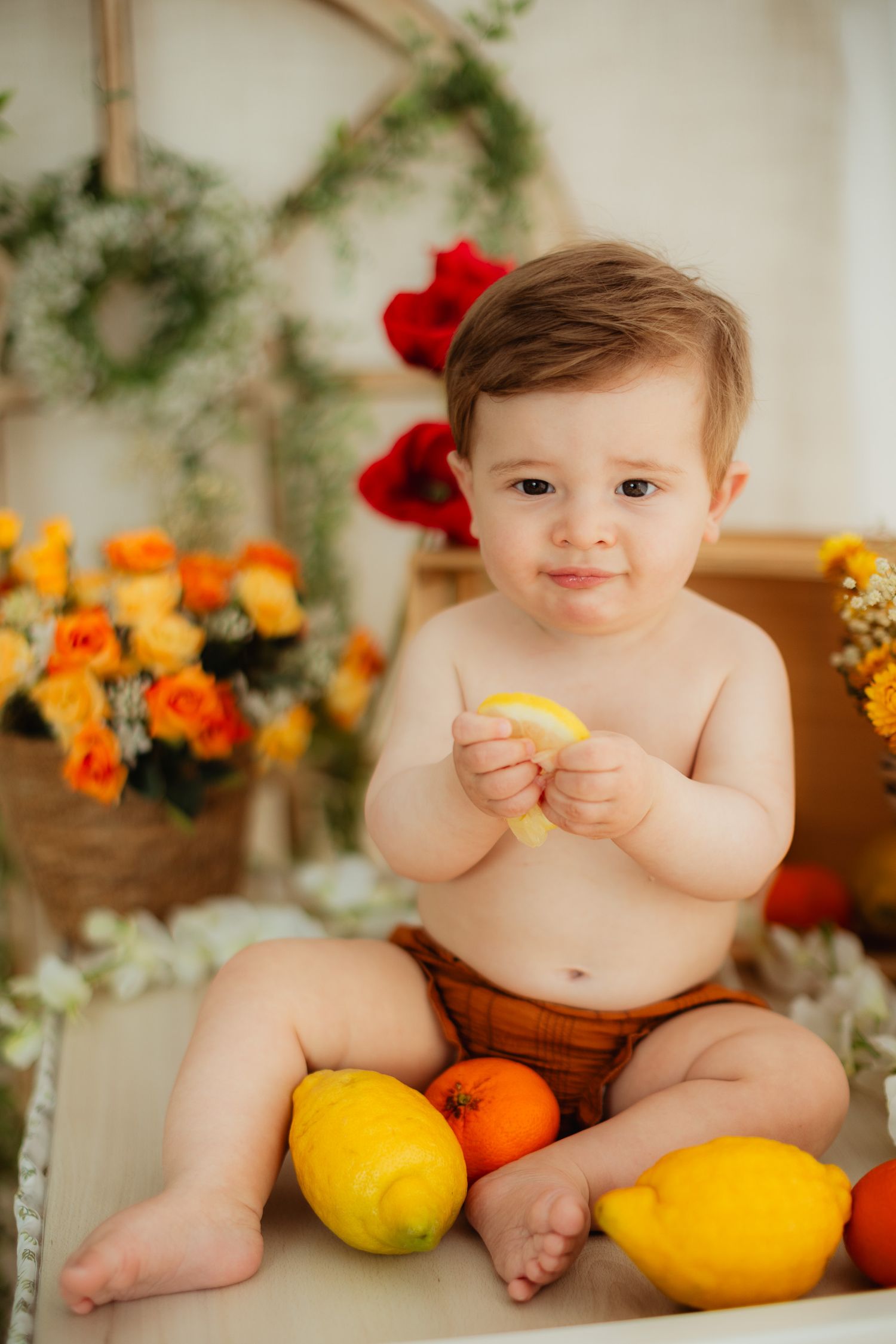 Cute baby boy sitting shirtless holding a lemon slice surrounded by fresh fruits and colorful flowers in a bright studio.