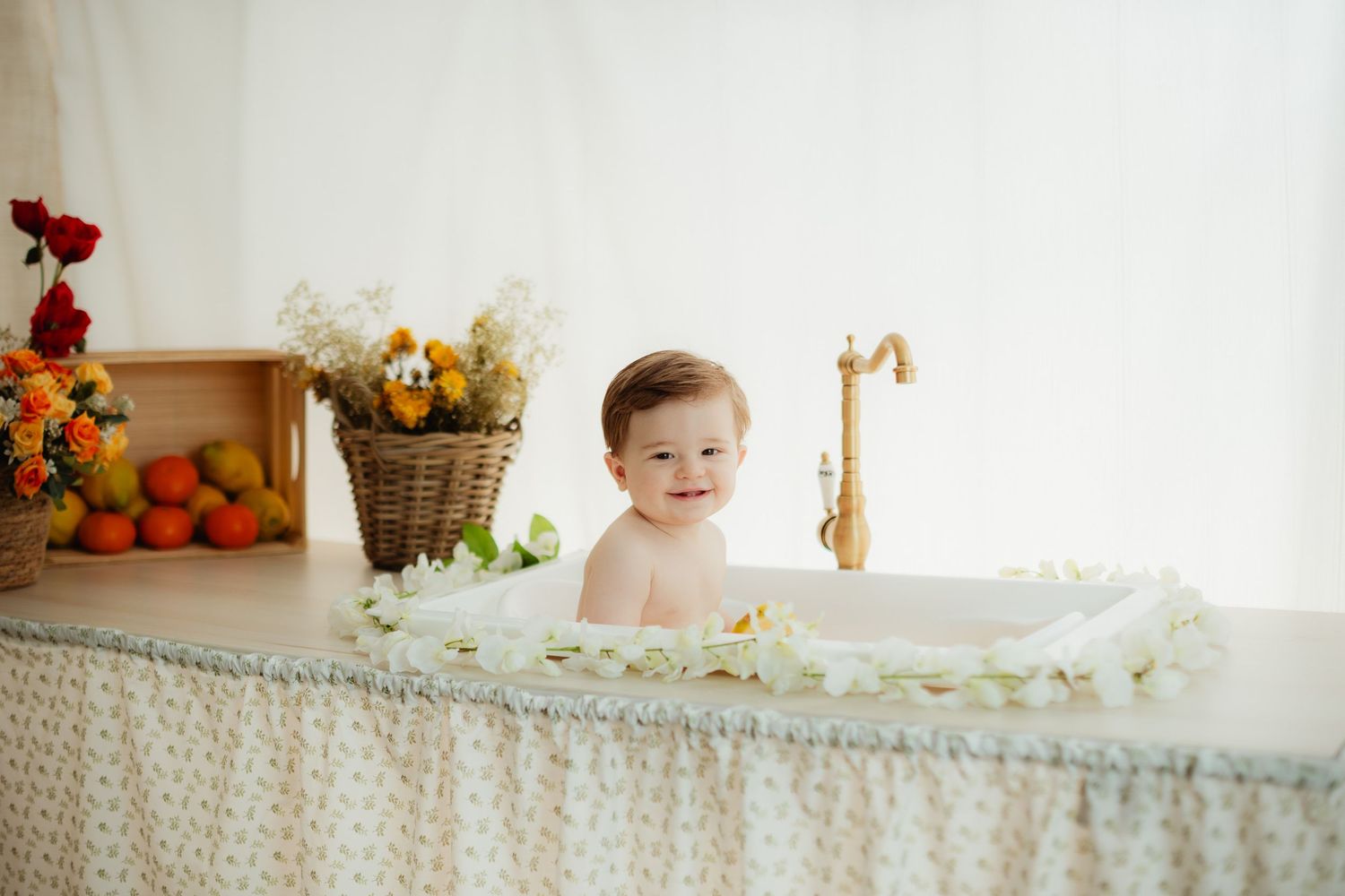 A happy baby sits in a floral milk bath surrounded by flower petals, with a basket of flowers and fruits nearby.