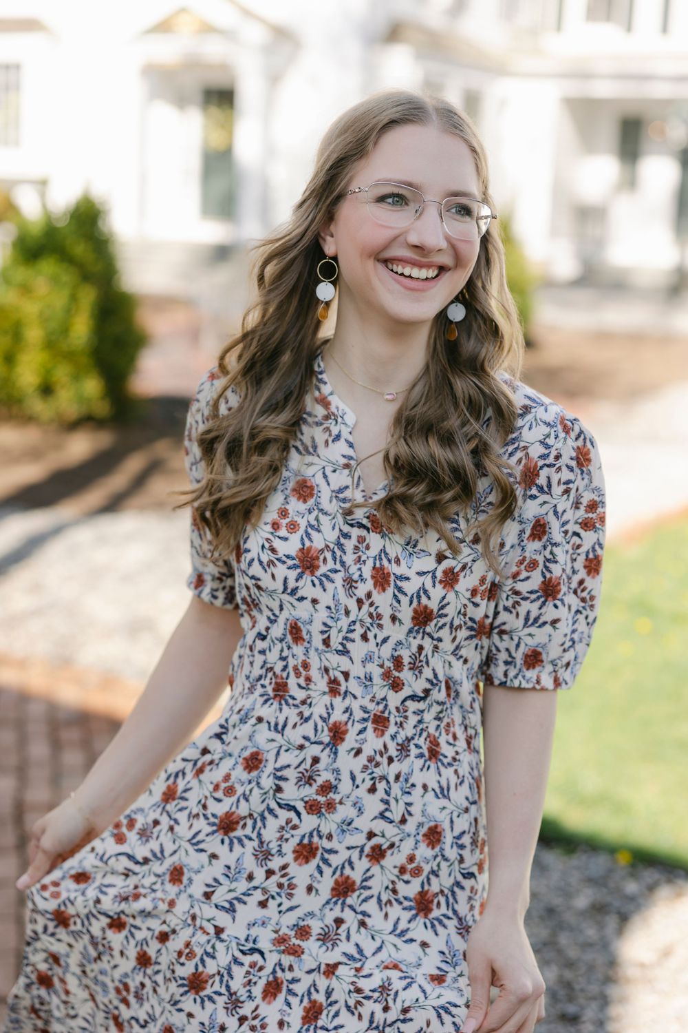 Smiling young woman in floral midi dress and glasses with dangling earrings outdoors. Essex, Connecticut.