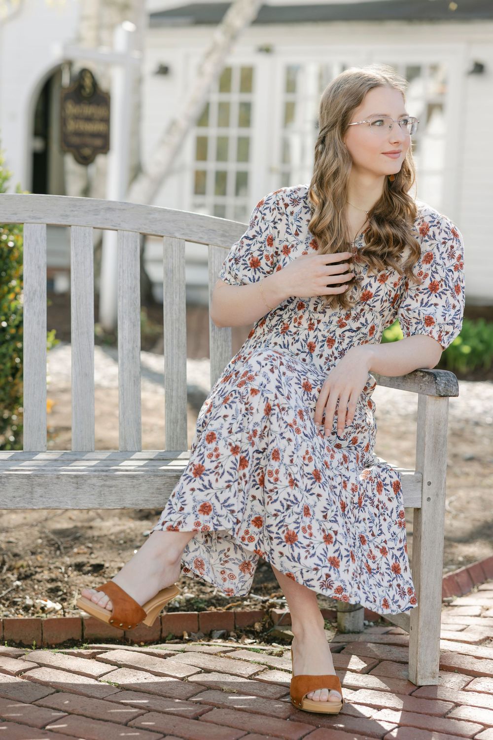Young woman in floral midi dress and tan sandals sitting on wooden bench outdoors. Essex, Connecticut.