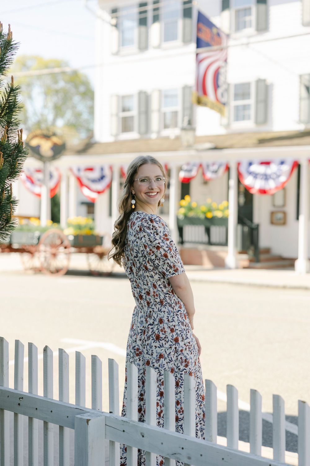Young woman in floral dress smiling by white picket fence with patriotic bunting decorations in background. Essex, Connecticut.