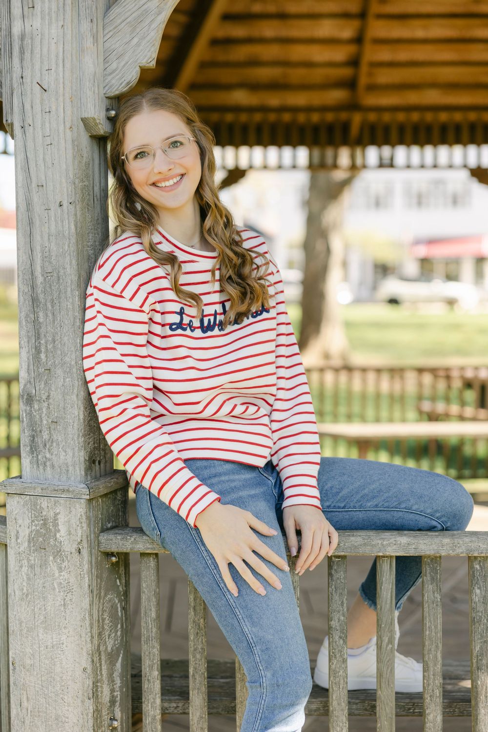 Young woman in red striped top and jeans smiling while seated on wooden railing under a gazebo outdoors. Essex, Connecticut.