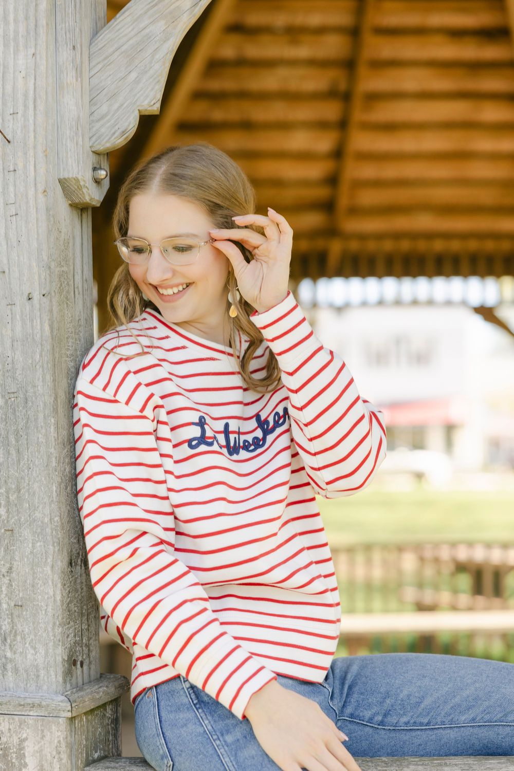 Smiling young girl in red striped shirt and jeans adjusting glasses, leaning against wooden gazebo outdoors. Essex, Connecticut.