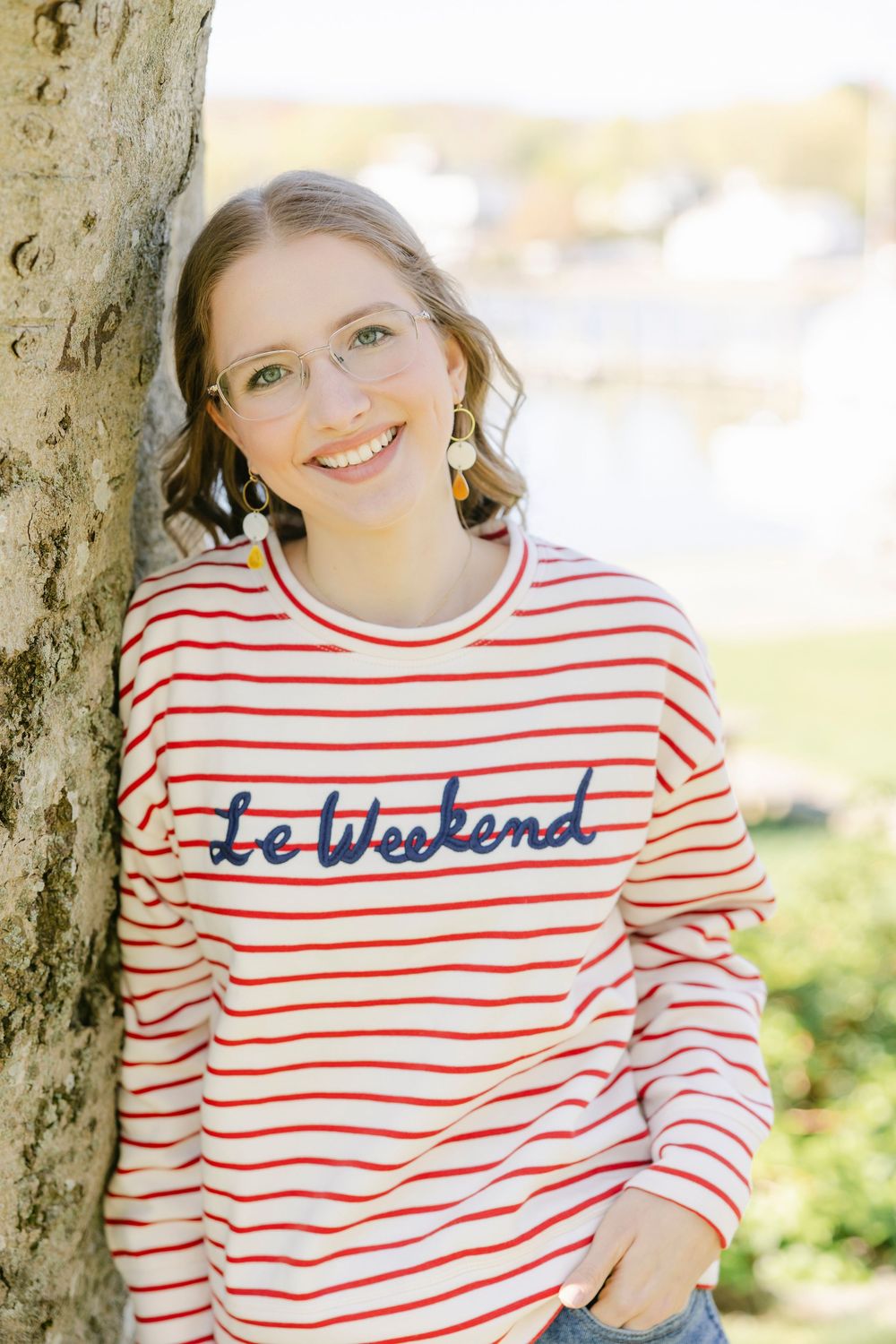 Smiling young woman in red striped Le Weekend sweatshirt leaning against tree outdoors. Essex, Connecticut.