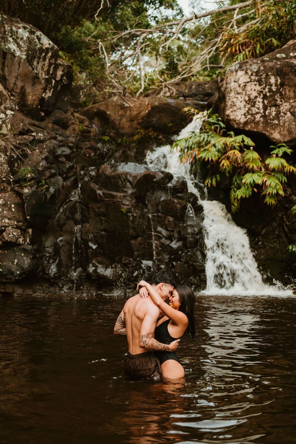Couple embracing and kissing in a natural pool before a lush tropical waterfall surrounded by rocks.