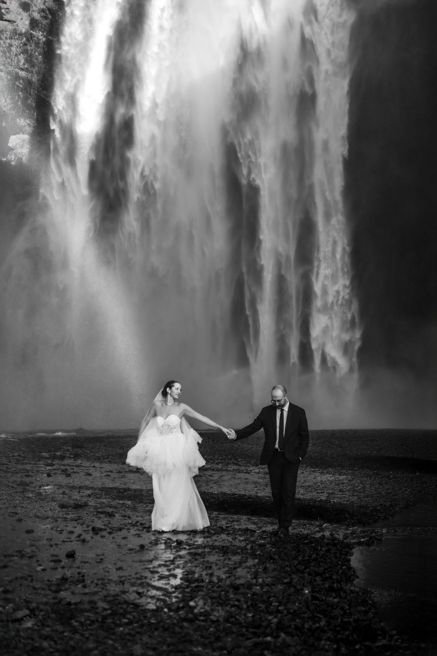 Black and white photo of bride and groom holding hands before a massive waterfall in Iceland.