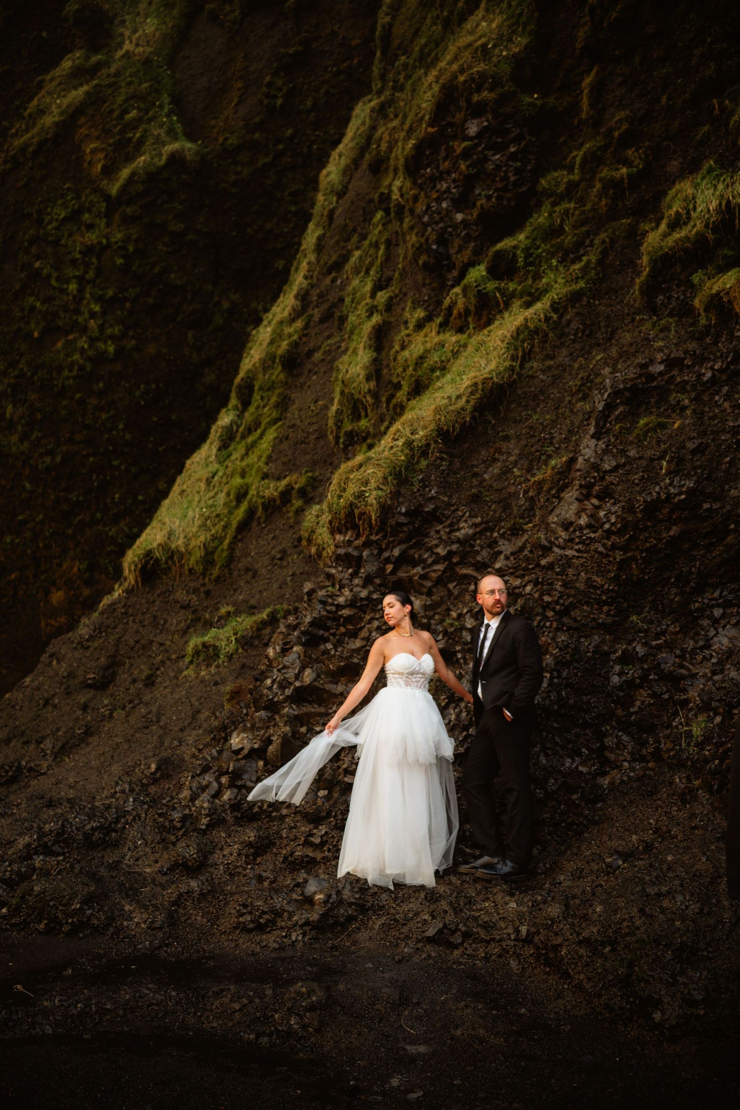 Bride and groom stand against mossy rocky cliffs in dramatic outdoor wedding portrait.