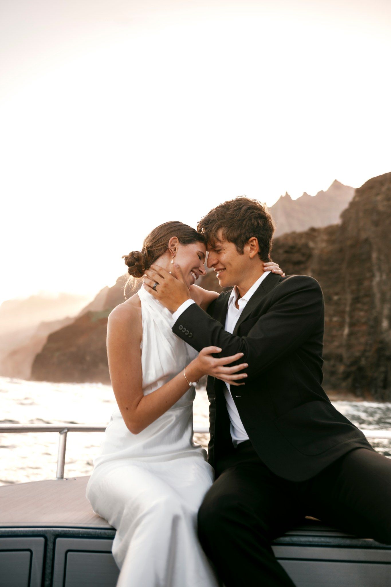 A bride and groom share a romantic moment on a yacht at sunset, with rocky cliffs and ocean waves in the background.