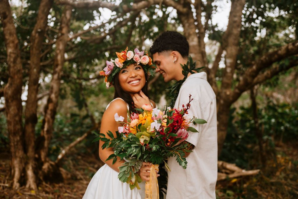 Couple in tropical forest, bride with floral crown holds colorful bouquet, groom in white shirt, sharing romantic moment.