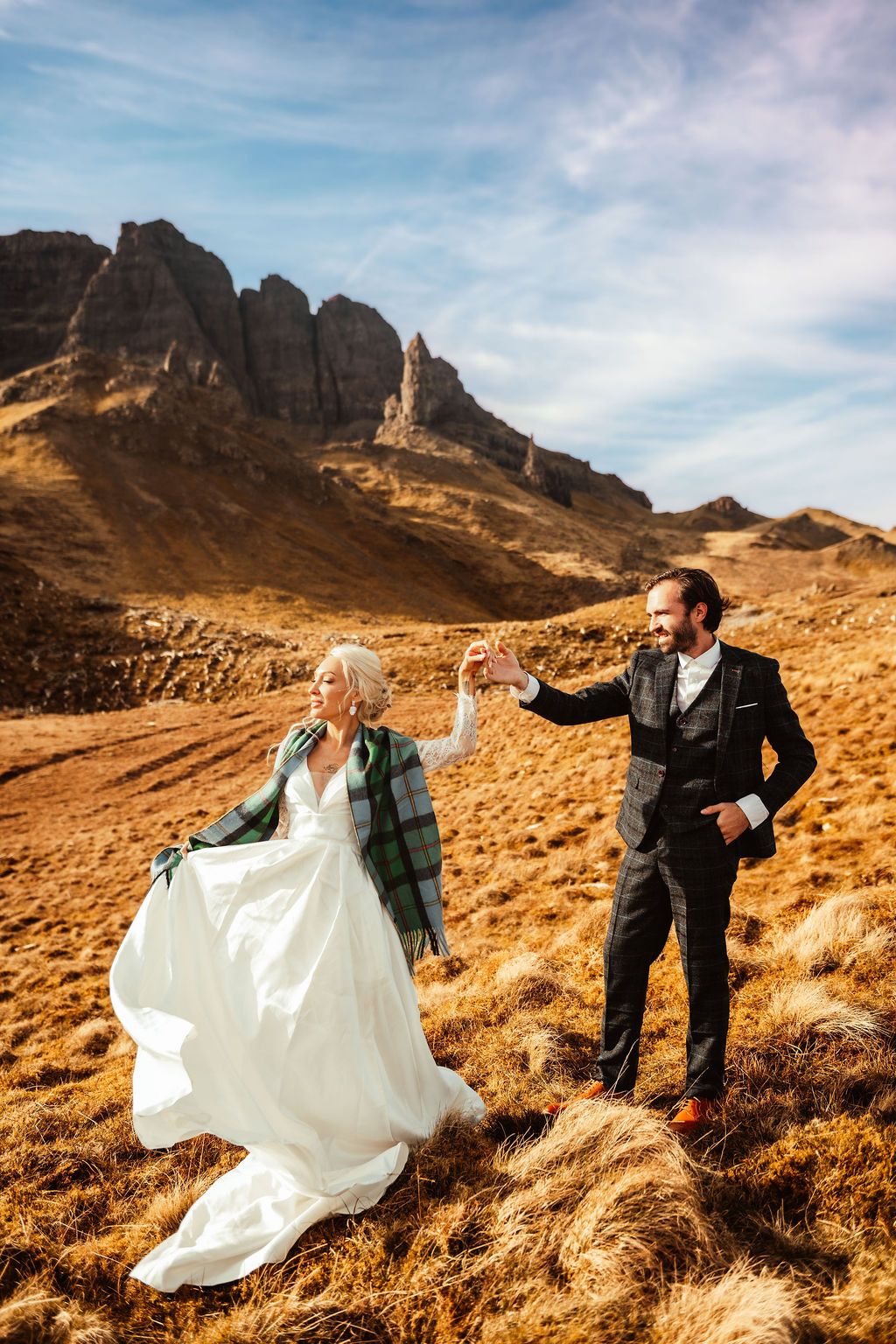 Bride and groom holding hands in Scottish Highlands, dramatic rocky peaks and golden moorland backdrop on their wedding day.