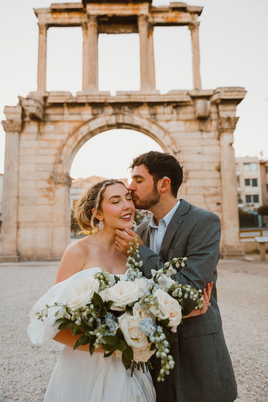 Couple in wedding attire kissing before Hadrian's Arch in Athens, bride holding white floral bouquet.