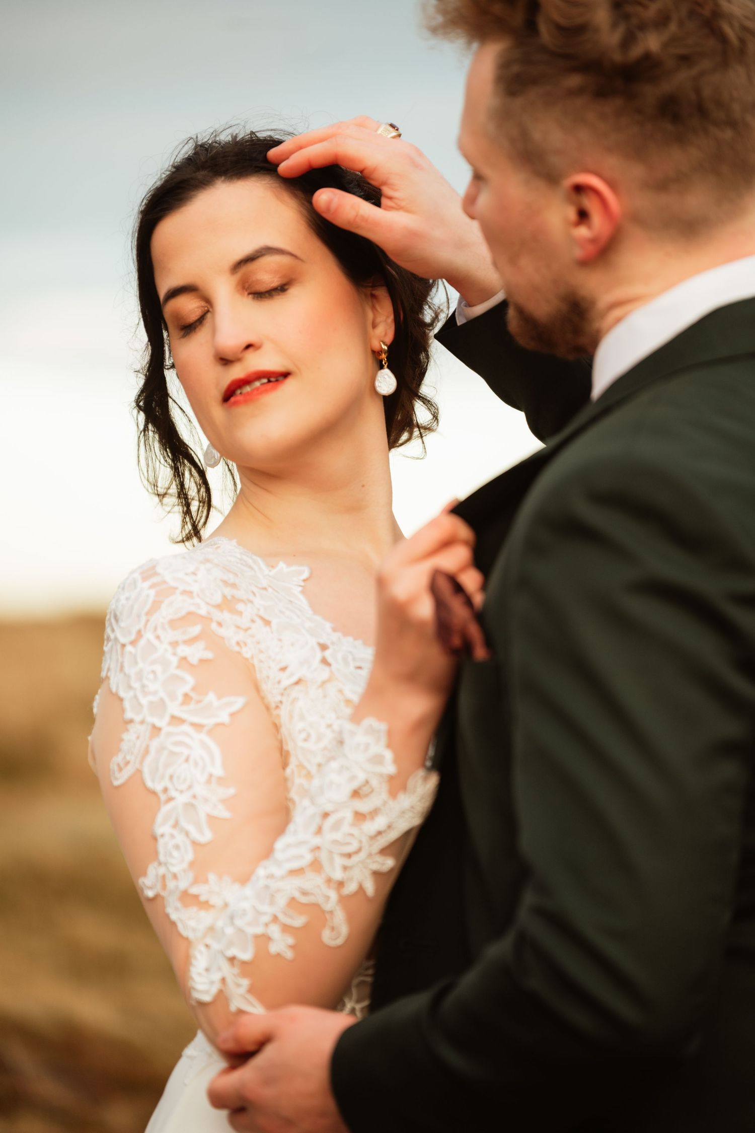 Groom tenderly touches brides hair as she closes her eyes in a romantic outdoor wedding portrait.