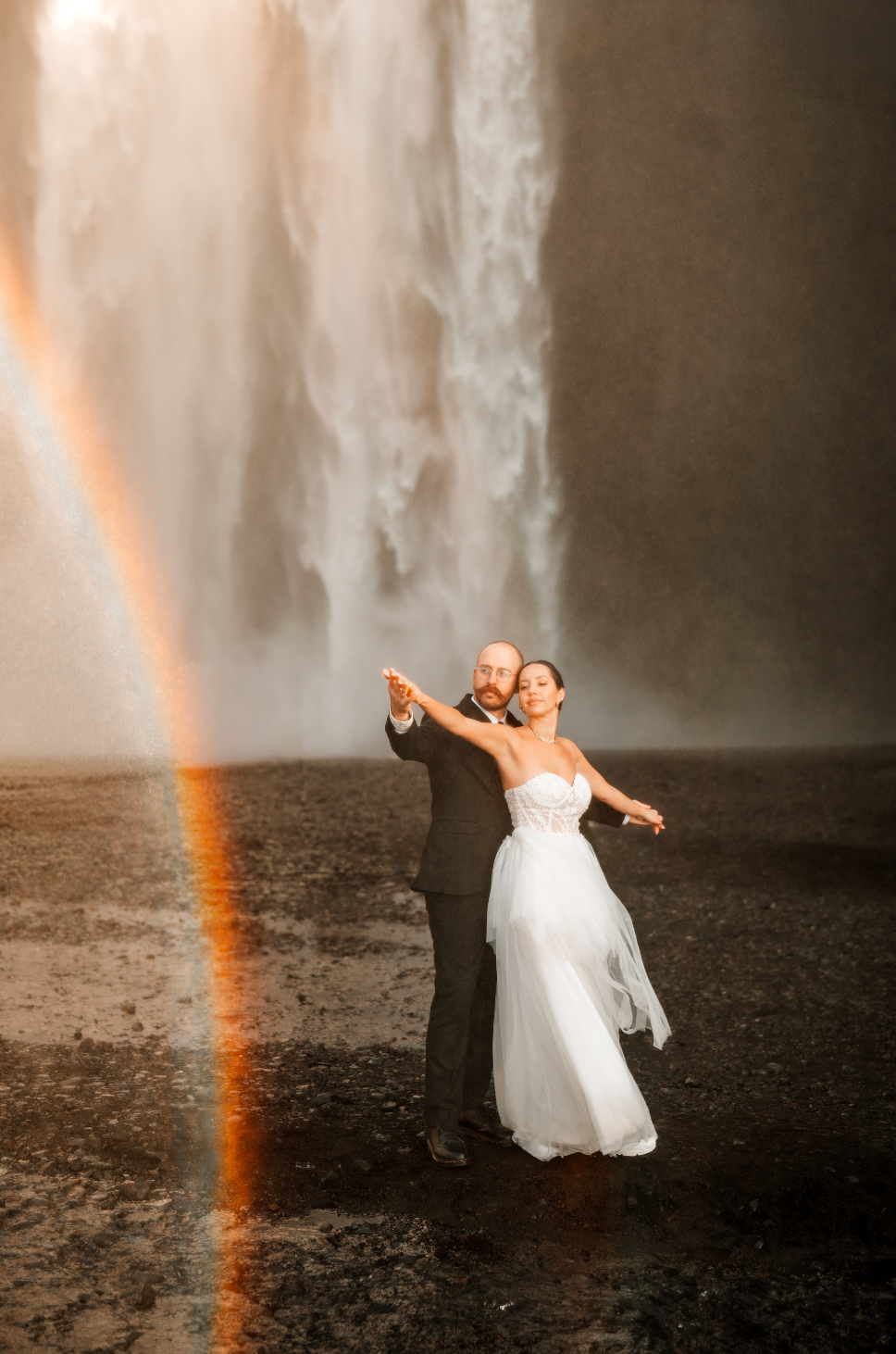 Couple dances before Skogafoss waterfall in Iceland with a vibrant rainbow arc during their elopement wedding.