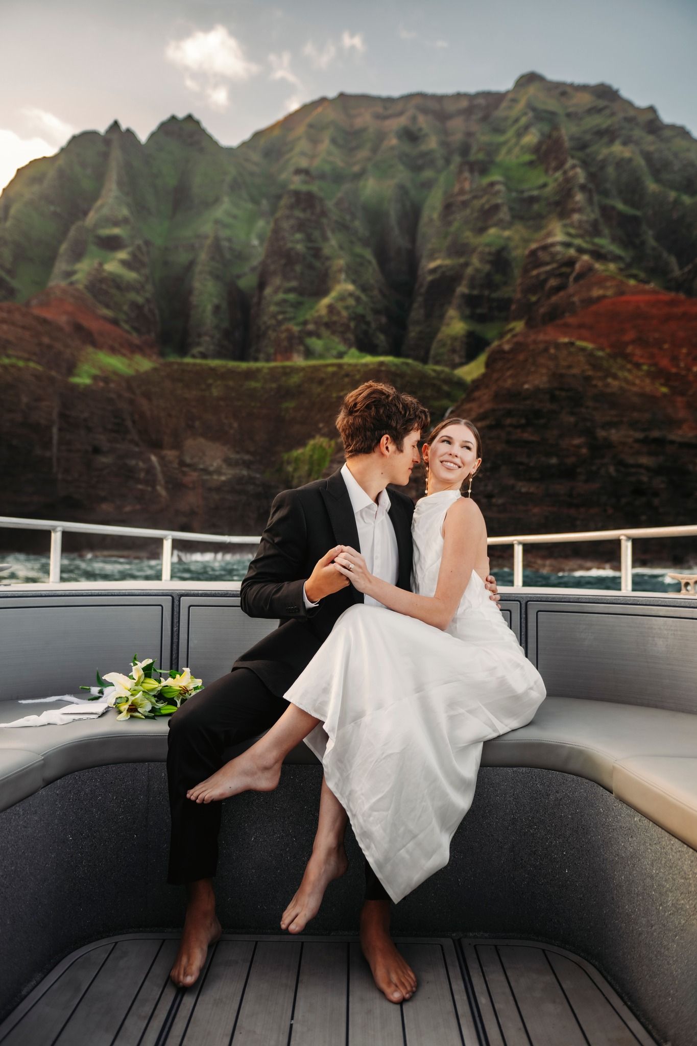 Bride and groom sitting barefoot on a yacht deck with dramatic Na Pali Coast cliffs in Hawaii behind them.