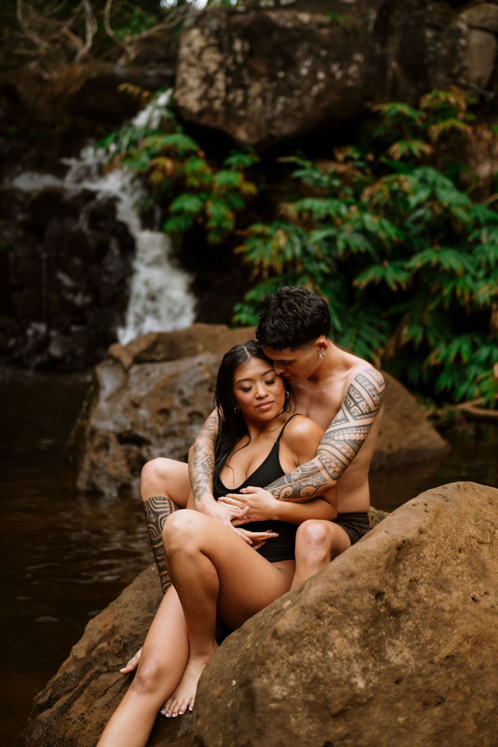 Couple sitting on rocks by a tropical waterfall, man with tattoos embracing woman in black swimsuit.
