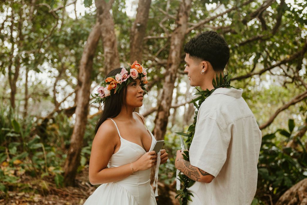 Couple in tropical forest, bride wearing floral crown and white dress, groom in white shirt with lei, sharing a romantic moment.